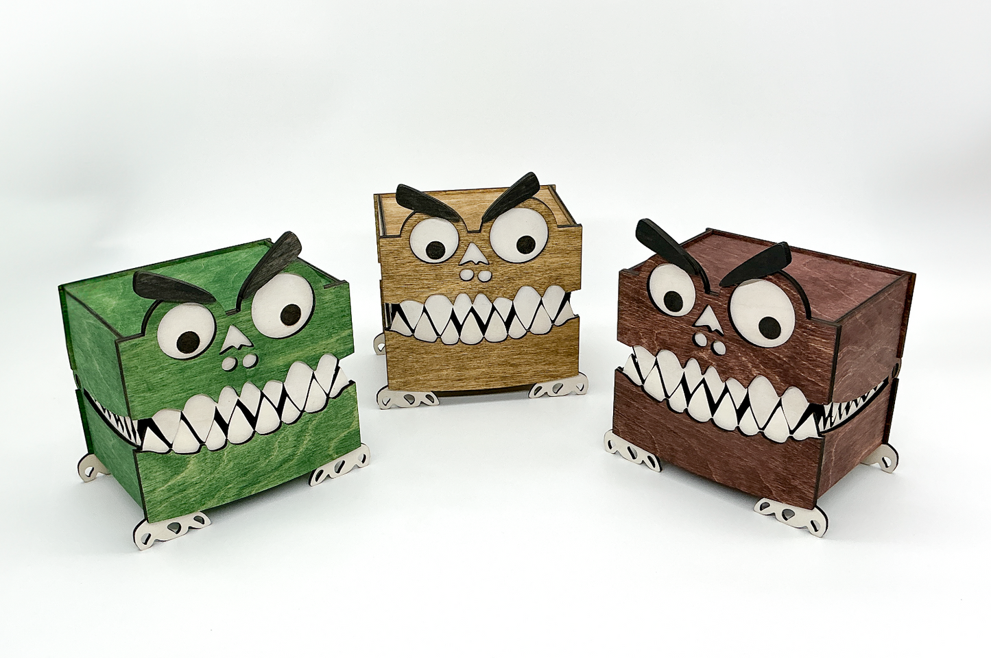 A trio of playful wooden monster storage boxes standing side-by-side. From left to right, one box is stained a rich mahogany, the center box a dark walnut, and the third a vibrant green. All three feature googly eyes, bushy eyebrows, white teeth, and small wooden feet, showcasing the different color options available.