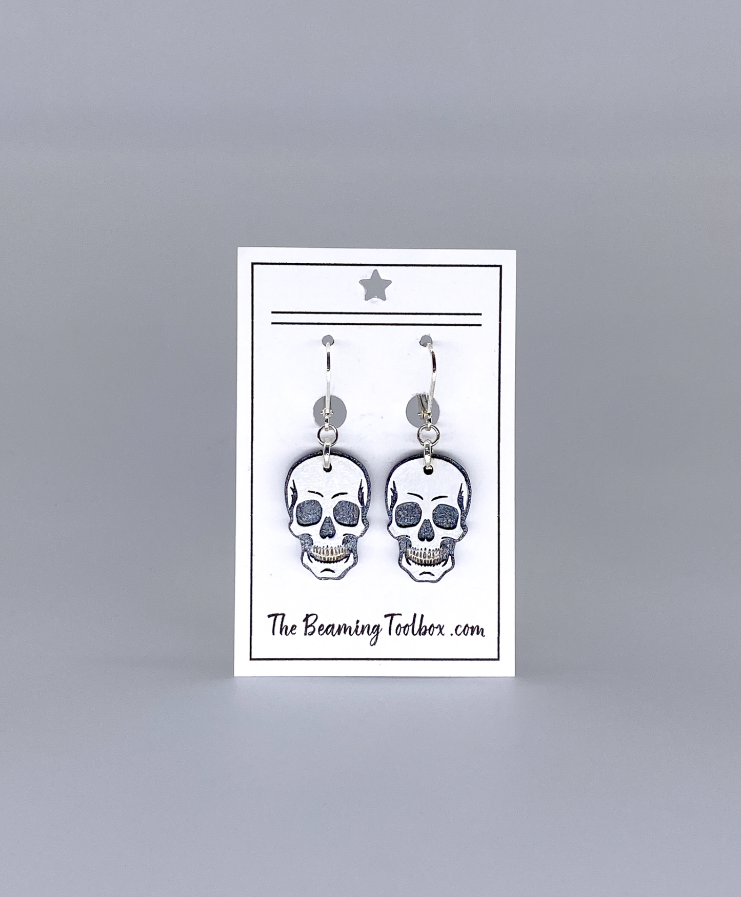 Skull-shaped earrings on a display card with 'The Beaming Toobox' branding against a gray background.