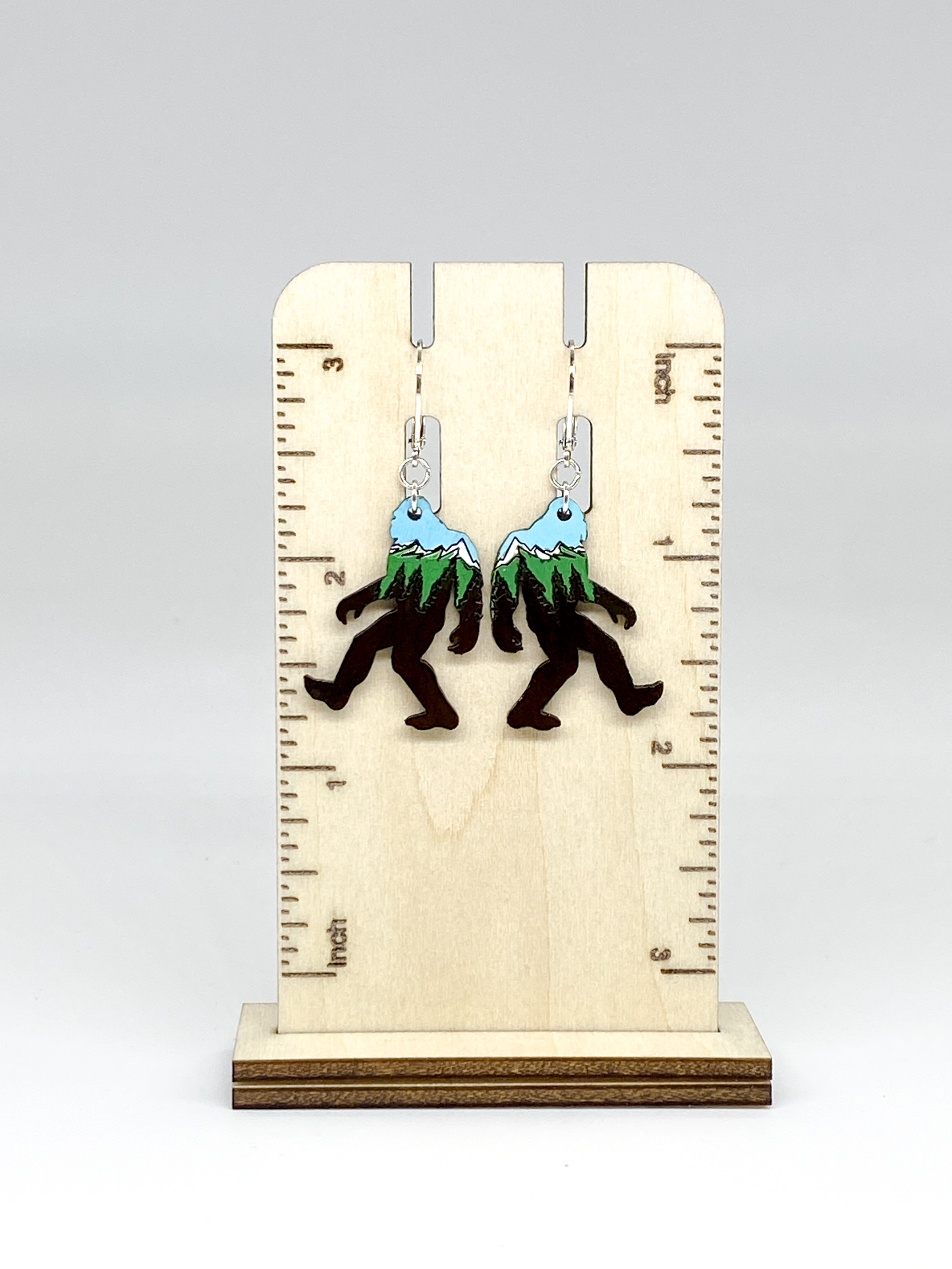 Sasquatch earrings with mountain design on a wooden stand with ruler for scale.