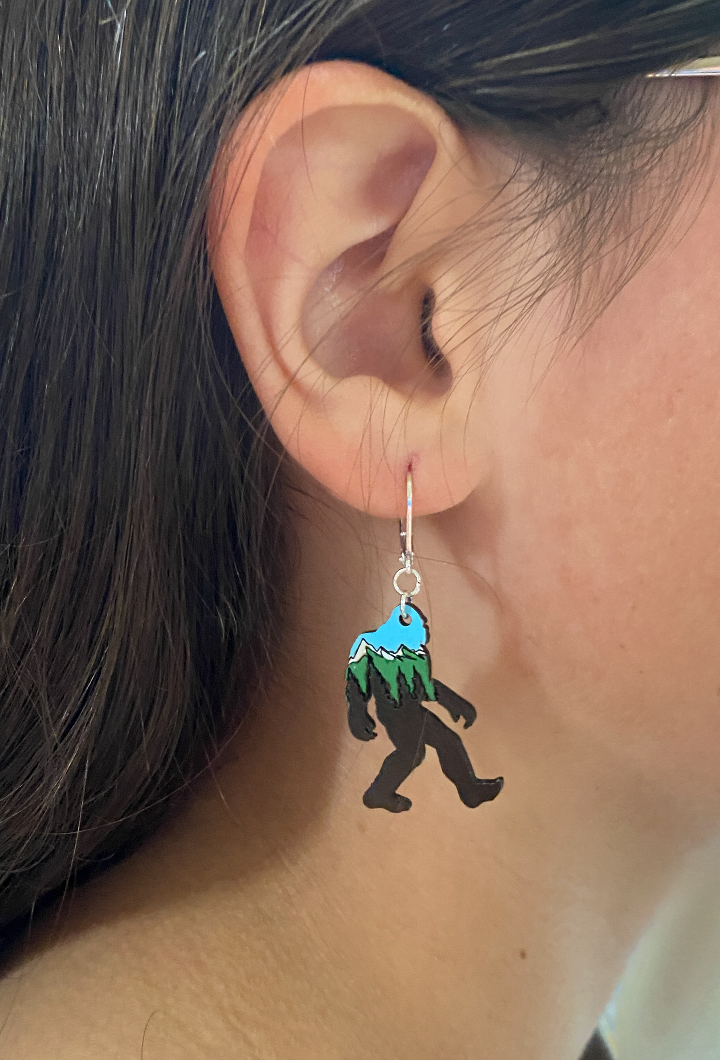 Earring shaped like a Sasquatch with a stylized mountain scene, worn by a person.
