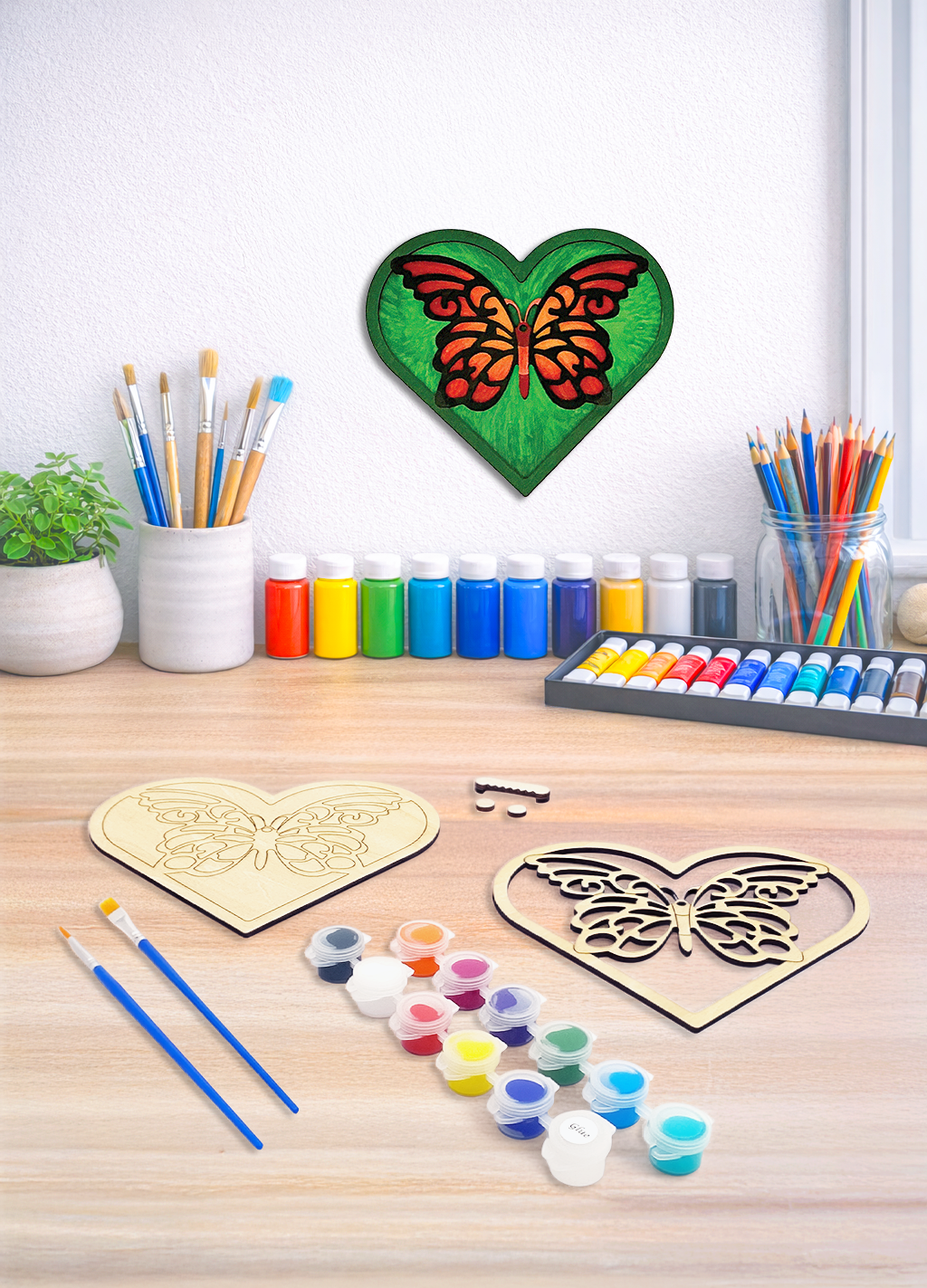 A DIY wood painting kit for a 3D layered butterfly heart featuring paint pots, brushes, and a finished version on a wall.