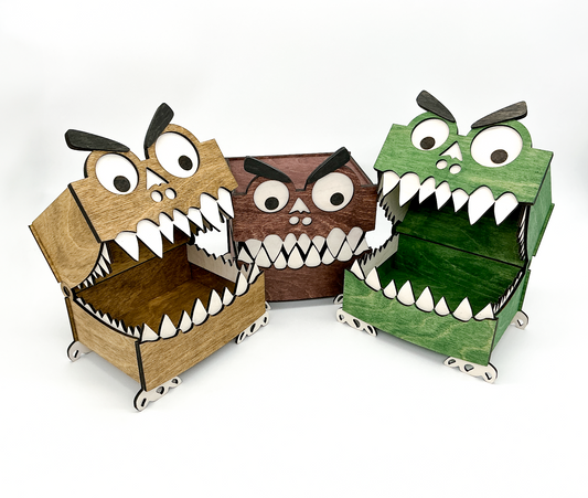 Three wooden Monster Boxes displayed side by side—one stained walnut, one mahogany, and one green. Two of the monsters have their hinged lids open, showing white teeth as if their mouths are open. All boxes feature googly eyes, big eyebrows, and small wooden feet, showcasing their playful and whimsical design.