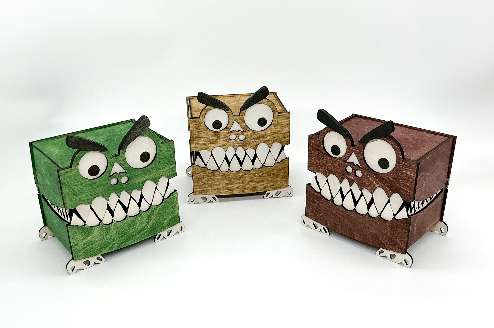 A trio of playful wooden monster storage boxes standing side-by-side. From left to right, one box is stained a rich mahogany, the center box a dark walnut, and the third a vibrant green. All three feature googly eyes, bushy eyebrows, white teeth, and small wooden feet, showcasing the different color options available.