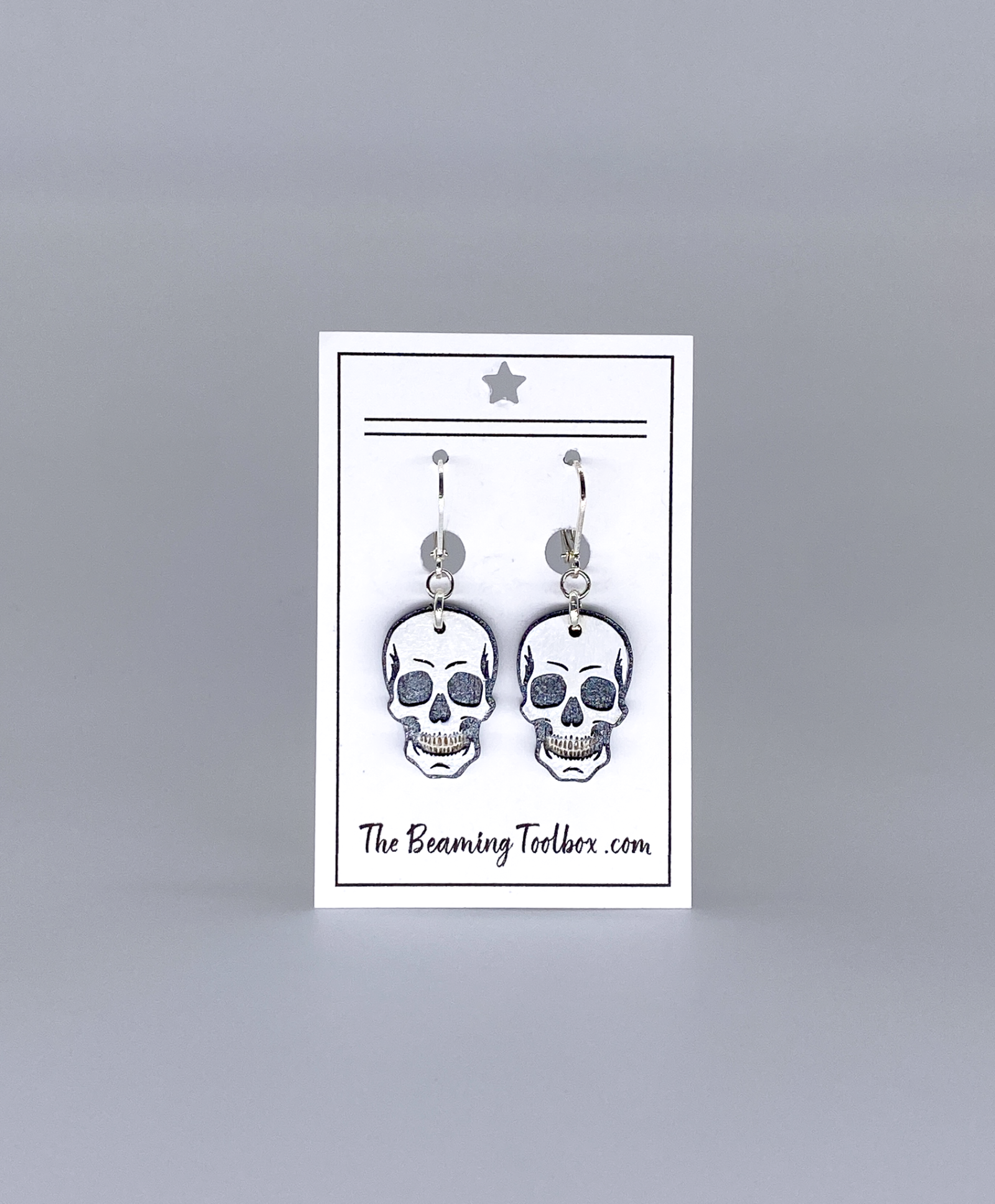 Skull-shaped earrings on a display card with 'The Beaming Toobox' branding against a gray background.