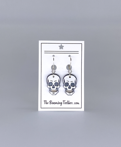 Skull-shaped earrings on a display card with 'The Beaming Toobox' branding against a gray background.