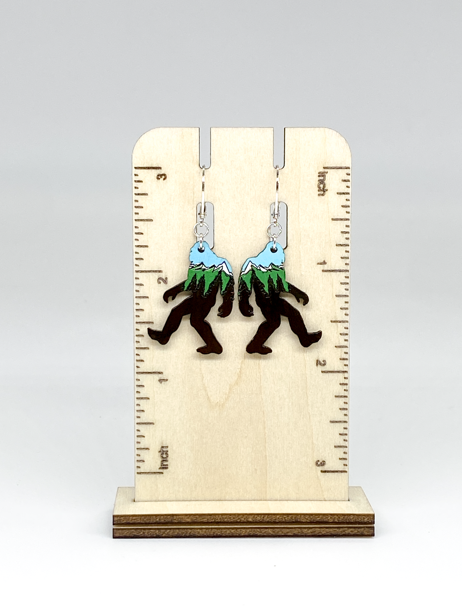 Sasquatch earrings with mountain design on a wooden stand with ruler for scale.