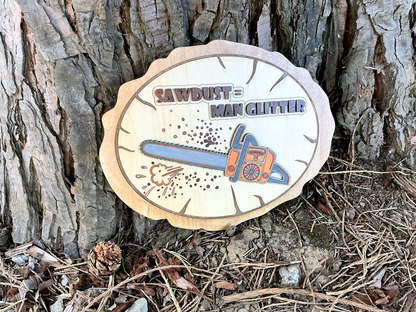 “Sawdust = Man Glitter” wooden chainsaw wall art displayed outdoors against a tree trunk in a rustic woodland setting.