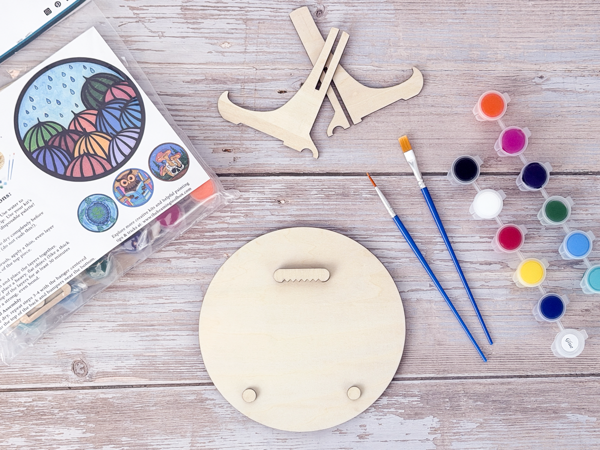 Umbrella adult craft kit supplies arranged with paint pots, brushes, display stand pieces, packaged kit, and blank wood back on a rustic wood background.