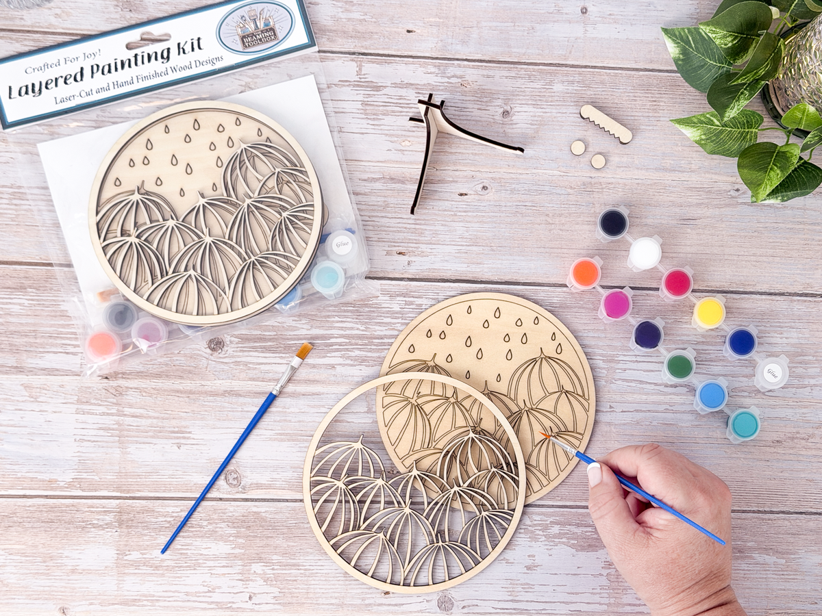 Umbrella DIY craft kit in progress with a hand painting the unpainted wood layers beside paint pots, brushes, packaged kit, and plant decor.