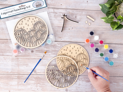 Umbrella DIY craft kit in progress with a hand painting the unpainted wood layers beside paint pots, brushes, packaged kit, and plant decor.