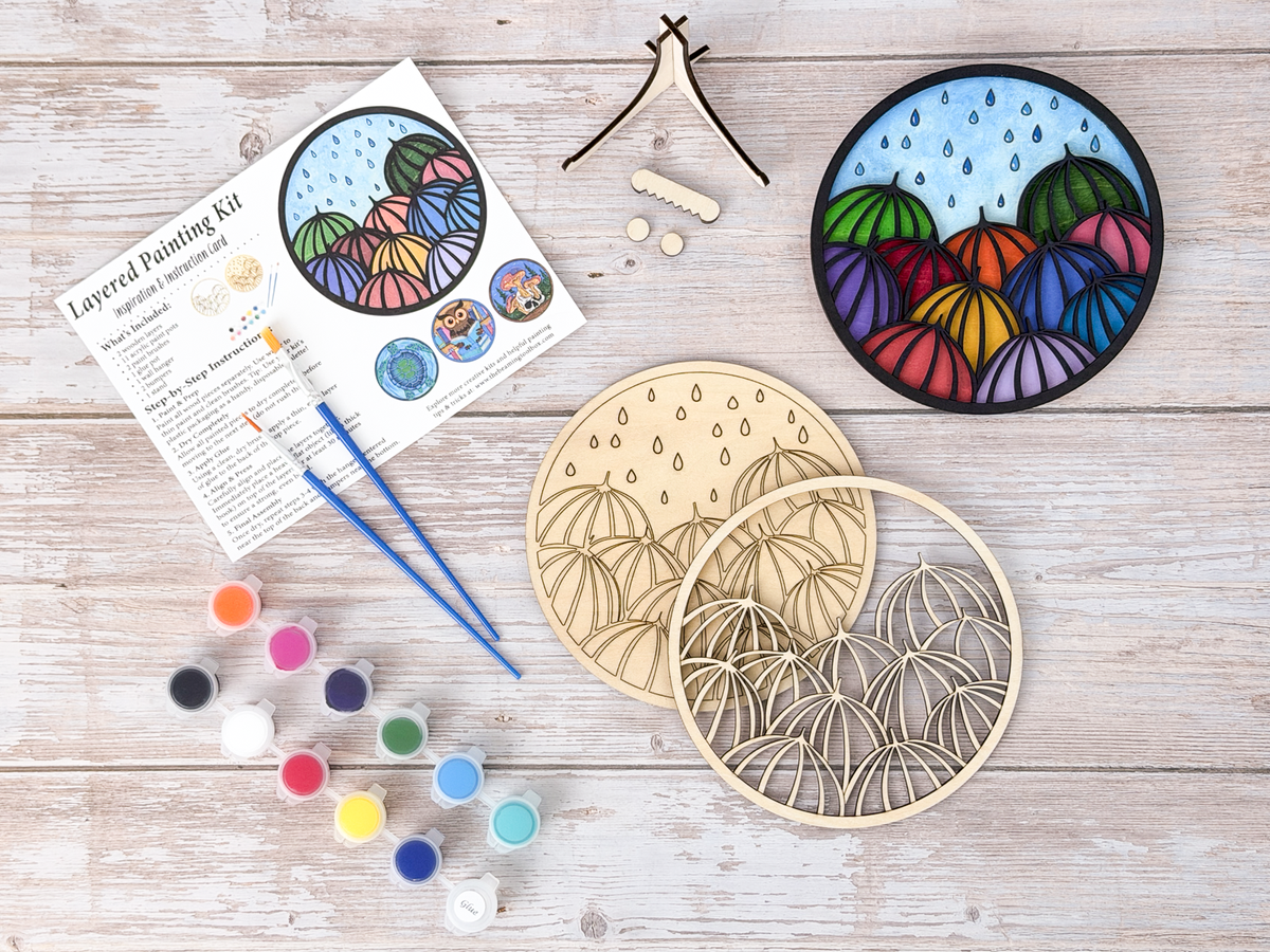 Umbrella painting kit with finished colorful umbrella artwork, unpainted wood layers, instruction card, paint pots, brushes, glue, and stand pieces.