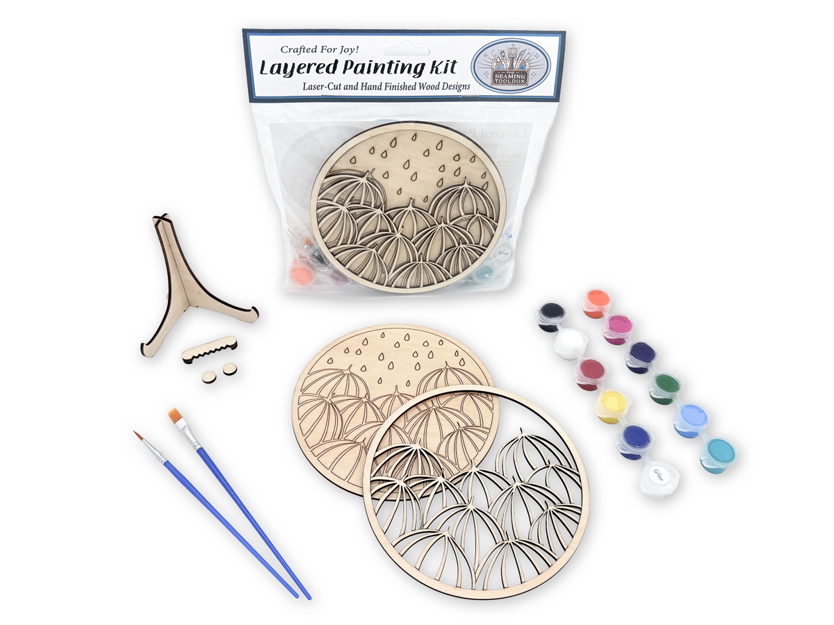 Umbrella painting kit for adults shown with packaged kit, unpainted layered wood pieces, paint pots, brushes, glue, and display stand.