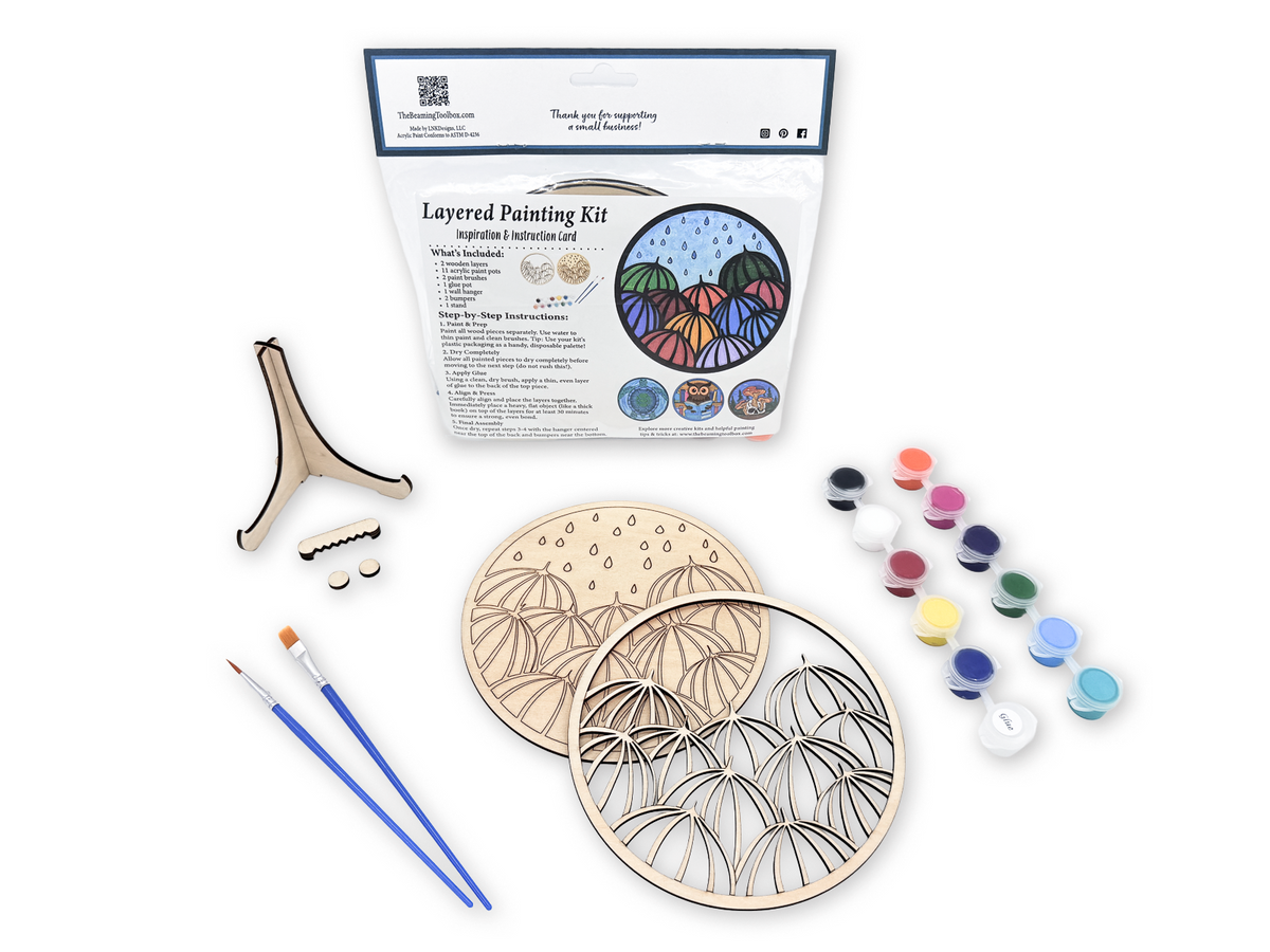 Unpainted umbrella relaxing adult craft kit arranged with paint pots, brushes, glue, display stand pieces, candle, and plant decor.