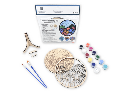 Unpainted umbrella relaxing adult craft kit arranged with paint pots, brushes, glue, display stand pieces, candle, and plant decor.
