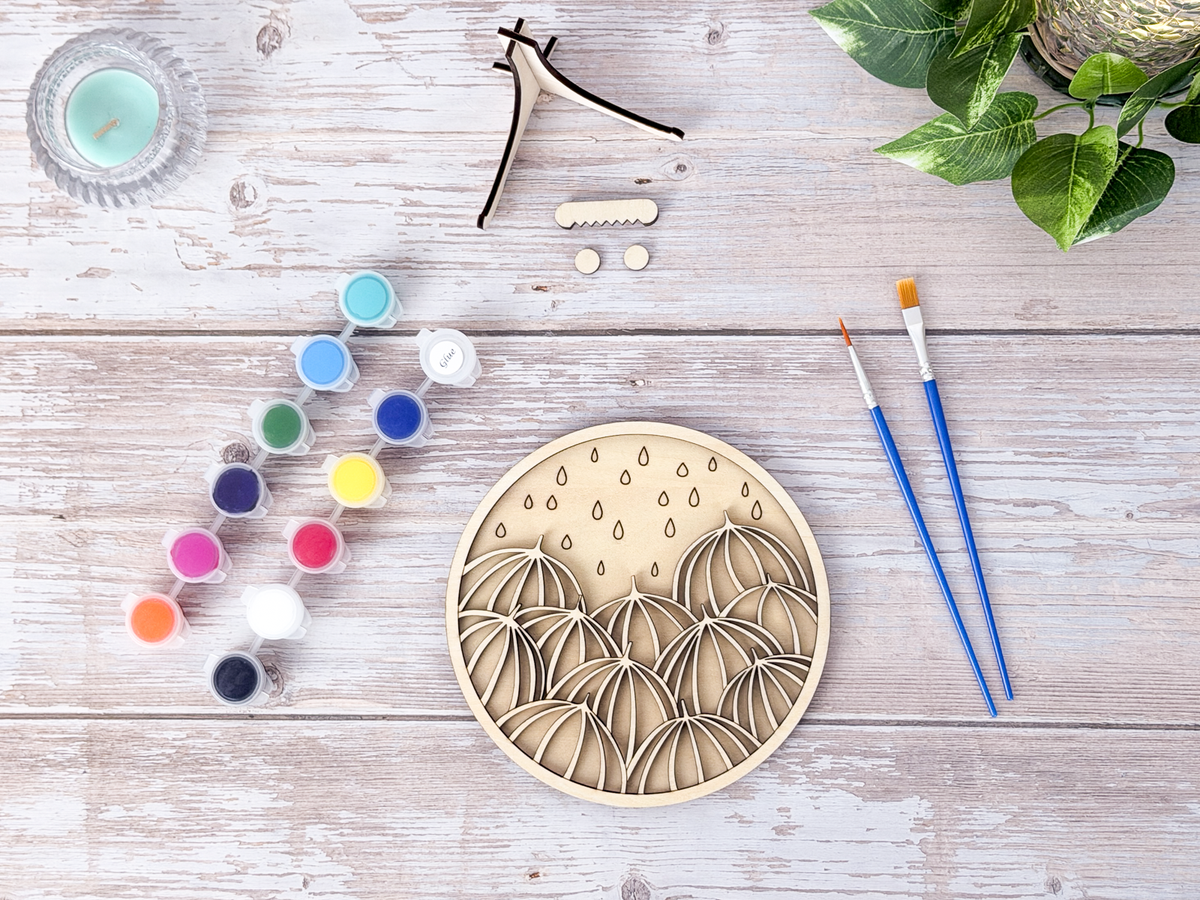 Umbrella unique wall art kit flat lay with unpainted wood layers, colorful finished example, instruction card, paint pots, brushes, glue, and stand pieces.