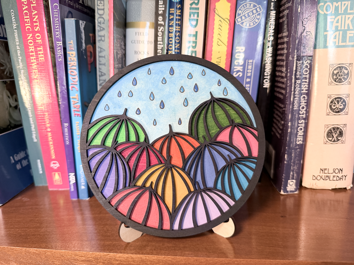 Finished umbrella layered wood painting displayed on a small stand with colorful umbrellas and blue raindrops in front of books.
