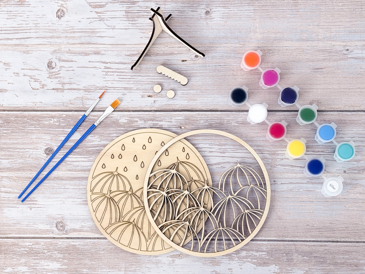Unpainted umbrella wood craft kit layers arranged with paint pots, blue brushes, glue, and display stand pieces on a rustic wood background.