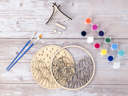 Unpainted umbrella wood craft kit layers arranged with paint pots, blue brushes, glue, and display stand pieces on a rustic wood background.
