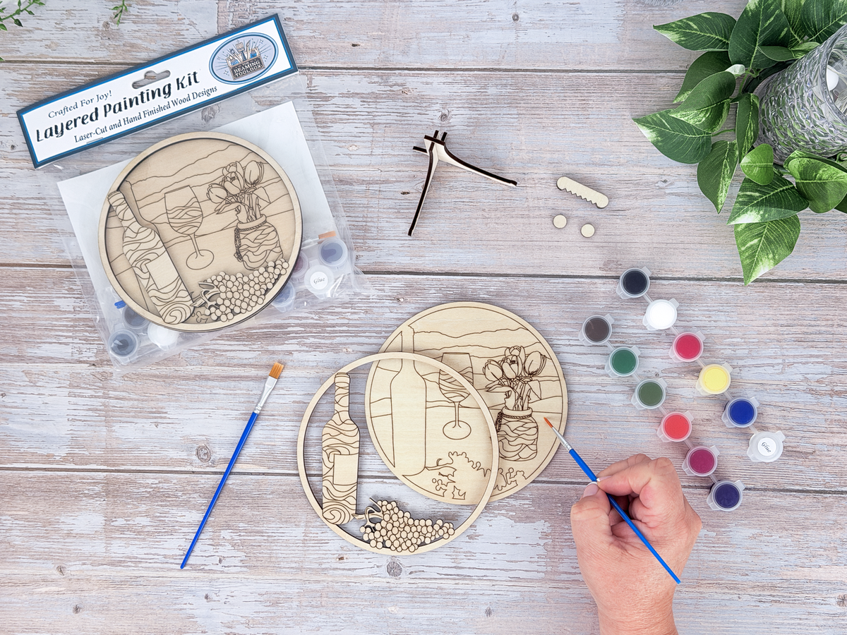 Wine and tulips paint and sip kit in progress with a hand painting the unpainted wood layers beside paint pots, brushes, packaged kit, and plant decor.