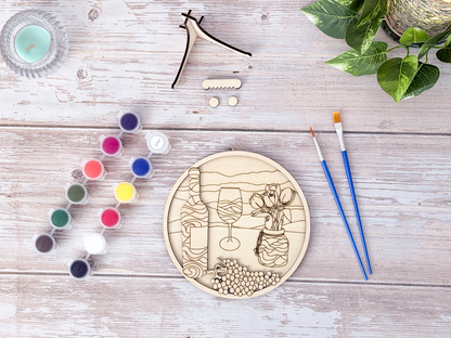 Unpainted wine and tulips relaxing craft kit arranged with paint pots, brushes, glue, display stand pieces, candle, and plant decor on a rustic wood background.