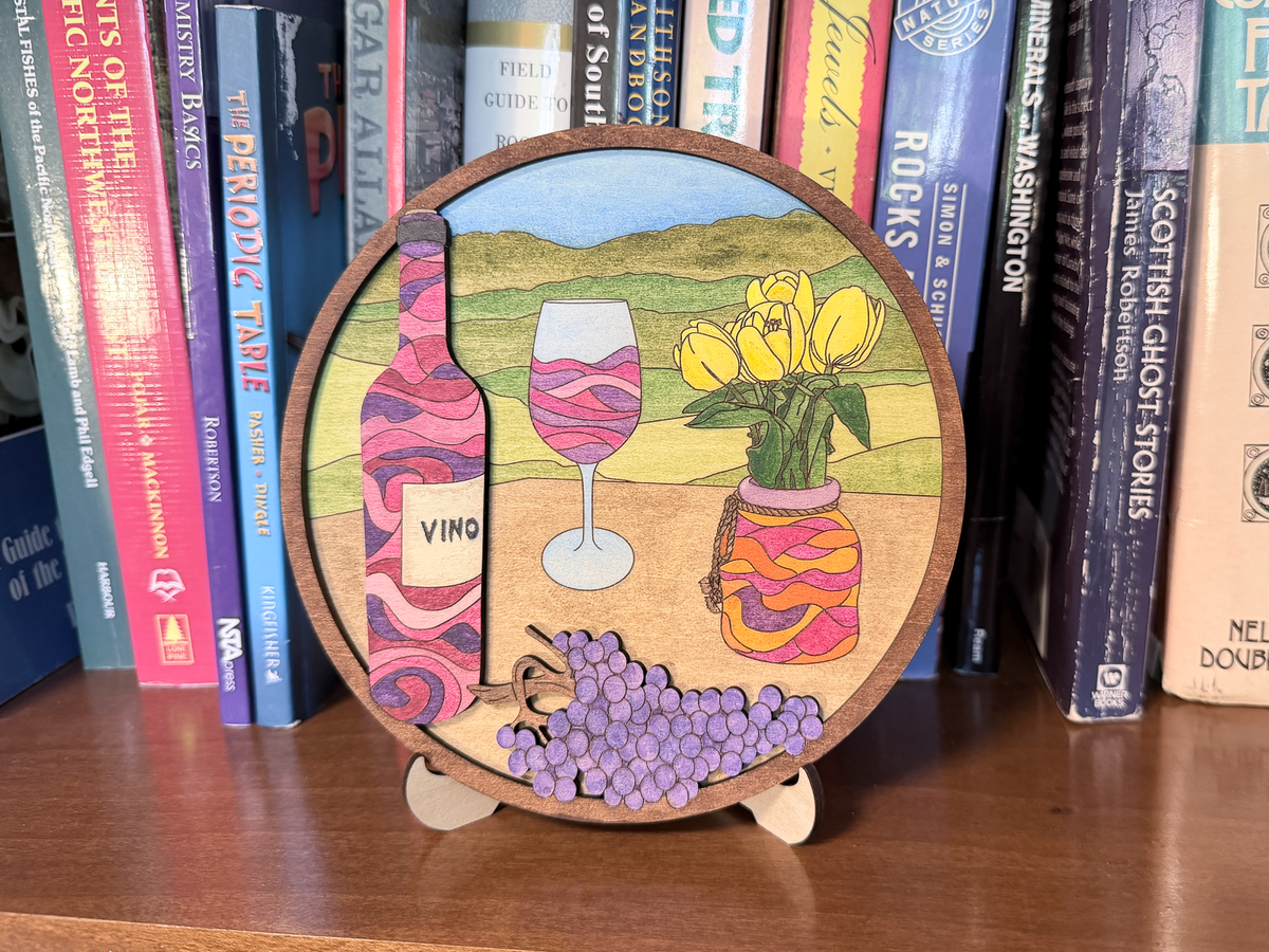 Finished wine and tulips layered wood painting displayed on a small stand with a painted wine bottle, wine glass, yellow tulips, purple grapes, and rolling hills.