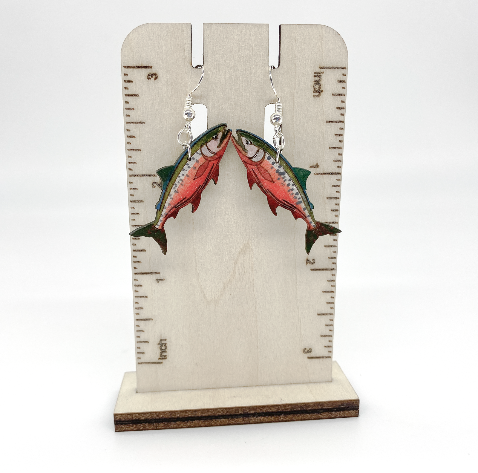 Salmon-shaped earrings on a wooden stand with measurement markings.