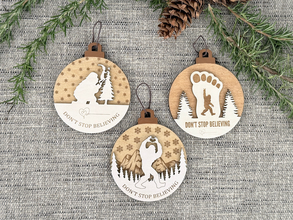Three wooden Sasquatch Christmas ornaments arranged on rustic fabric with evergreen branches and pinecones, showing three Don't Stop Believing designs.