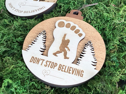 Wooden Sasquatch Christmas ornament with a large white Bigfoot footprint, walking Sasquatch silhouette, pine trees, and Don't Stop Believing engraving.