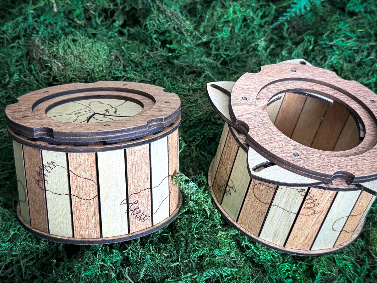 Two handcrafted wooden Bigfoot keepsake boxes on moss, showing the closed Sasquatch lid and an open box with its circular storage compartment.