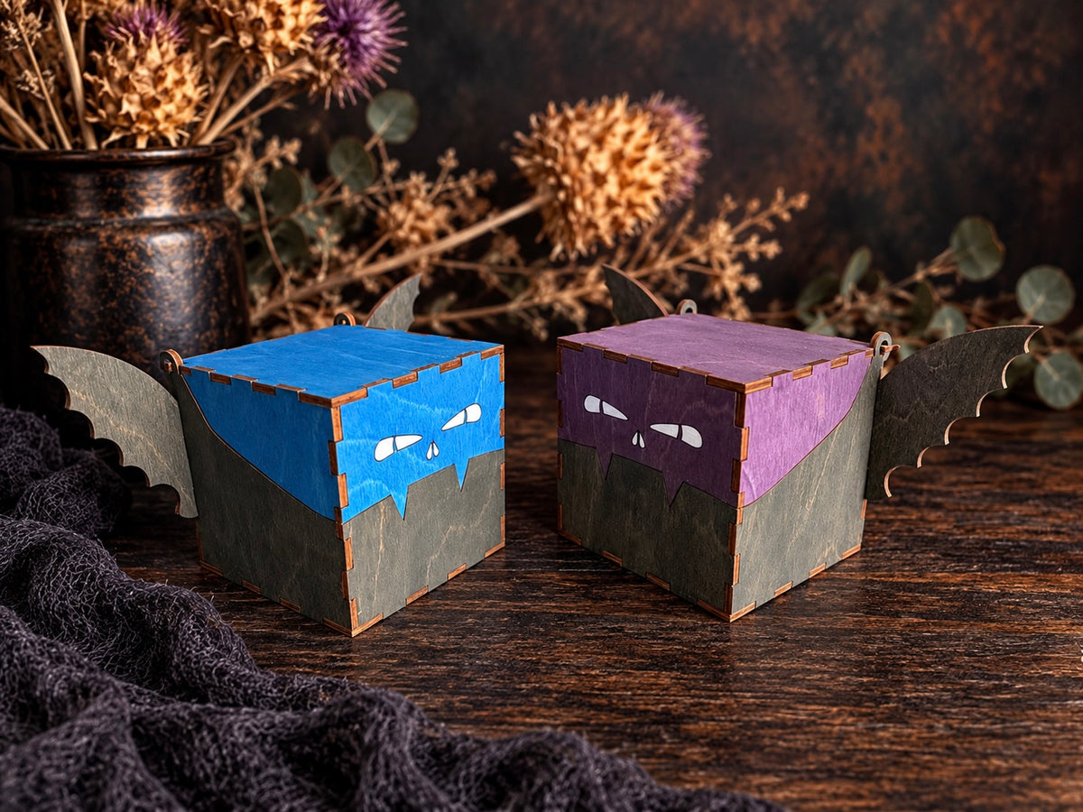Blue and purple handmade wooden bat jewelry boxes shown at an angle, highlighting their hinged lids, engraved faces, dark stained wings, and laser-cut wood joints.