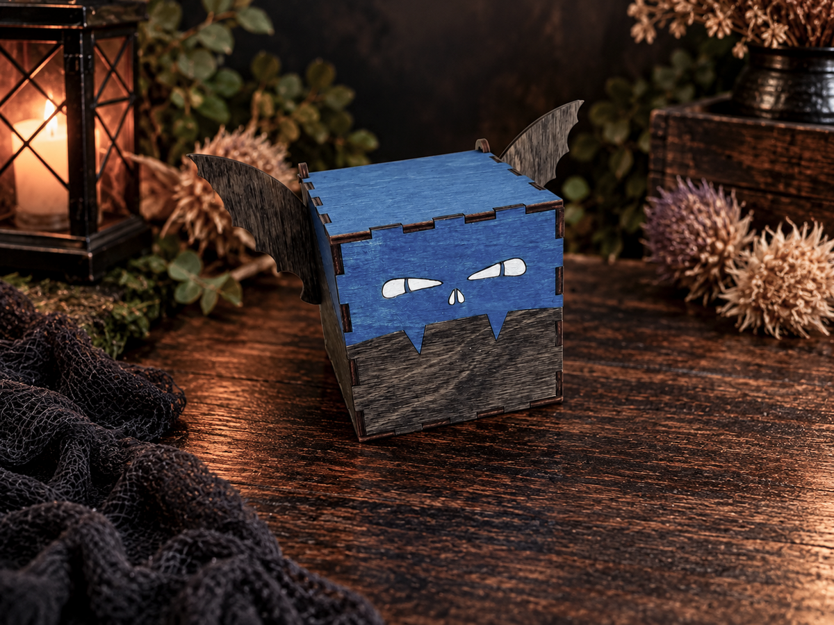 Handmade blue wooden bat trinket box with black wings, engraved white eyes, and hinged lid displayed on a rustic wooden table with Halloween lantern and greenery.