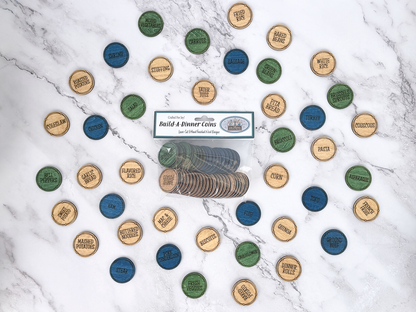 A set of round wooden game tokens labeled with food categories like pasta, rice, beans, and vegetables, arranged on a white marble background around a packaged version of the product that reads 'Build A Dinner Coins'.