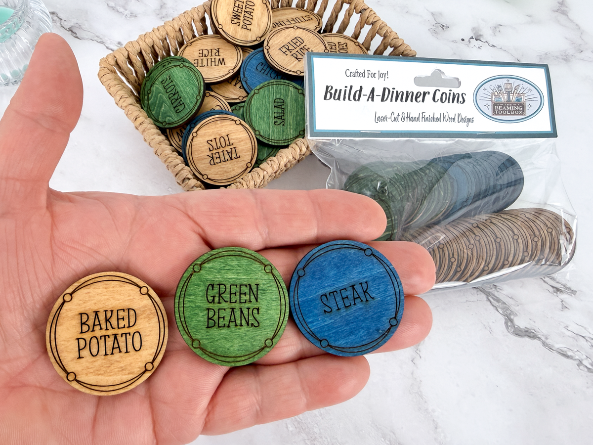 A hand holds three round wooden coin tokens labeled 'BAKED POTATO,' 'GREEN BEANS,' and 'STEAK,' with a basket and clear package of matching tokens behind them. The package card reads 'Build-A-Dinner Coins' and shows the brand 'The Beaming Toolbox'.