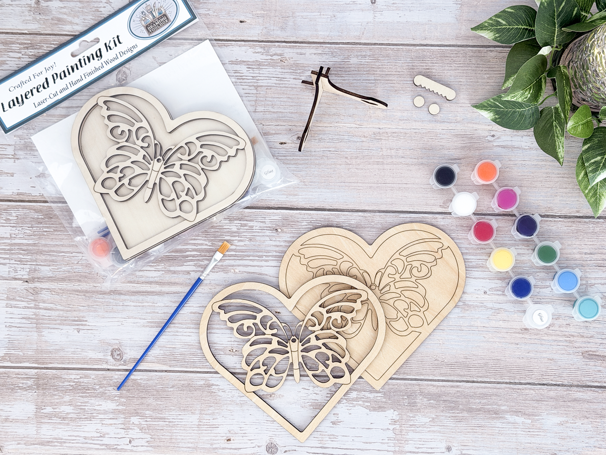 Unfinished butterfly heart wood craft kit pieces arranged with paint pots, brushes, display hardware, and packaging.