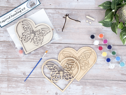 Unfinished butterfly heart wood craft kit pieces arranged with paint pots, brushes, display hardware, and packaging.