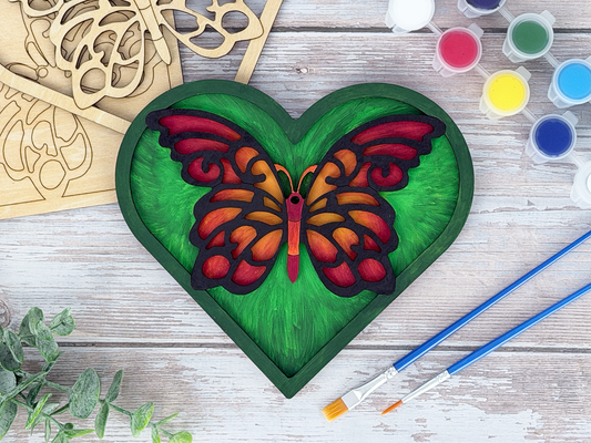Finished butterfly heart adult craft kit shown with painted layered wood artwork, unpainted kit pieces, paint pots, and brushes on a rustic wood background.