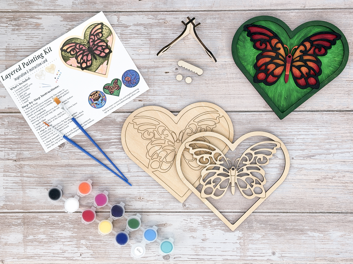 Complete butterfly heart adult craft kit with layered wood artwork, paint pots, brushes, glue, stand pieces, and packaged guide.