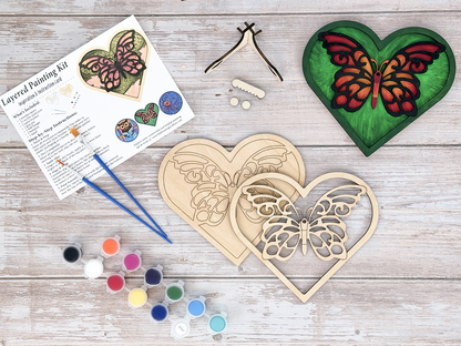 Complete butterfly heart adult craft kit with layered wood artwork, paint pots, brushes, glue, stand pieces, and packaged guide.
