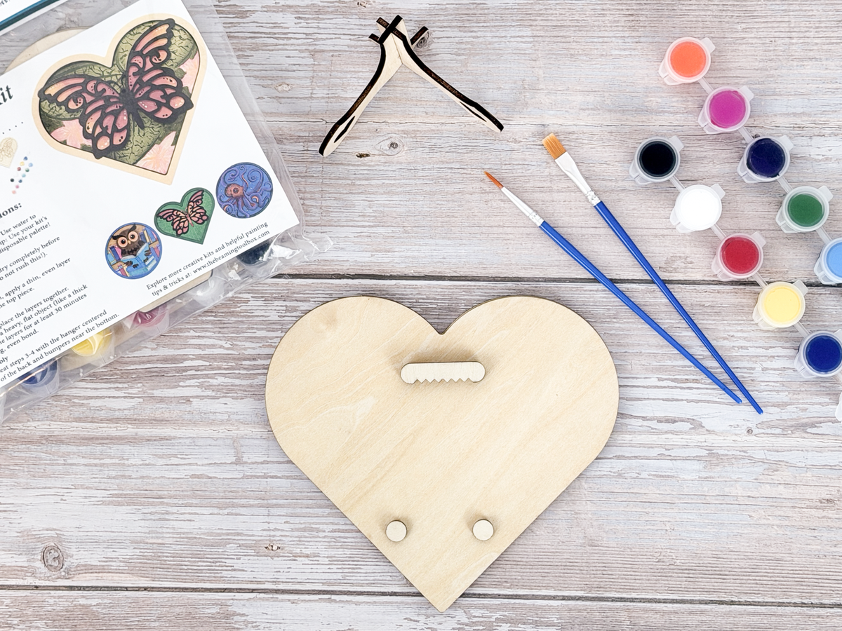 Back of the butterfly heart wood kit showing the hanger piece, small bumpers, brushes, paint pots, and packaged instruction card.