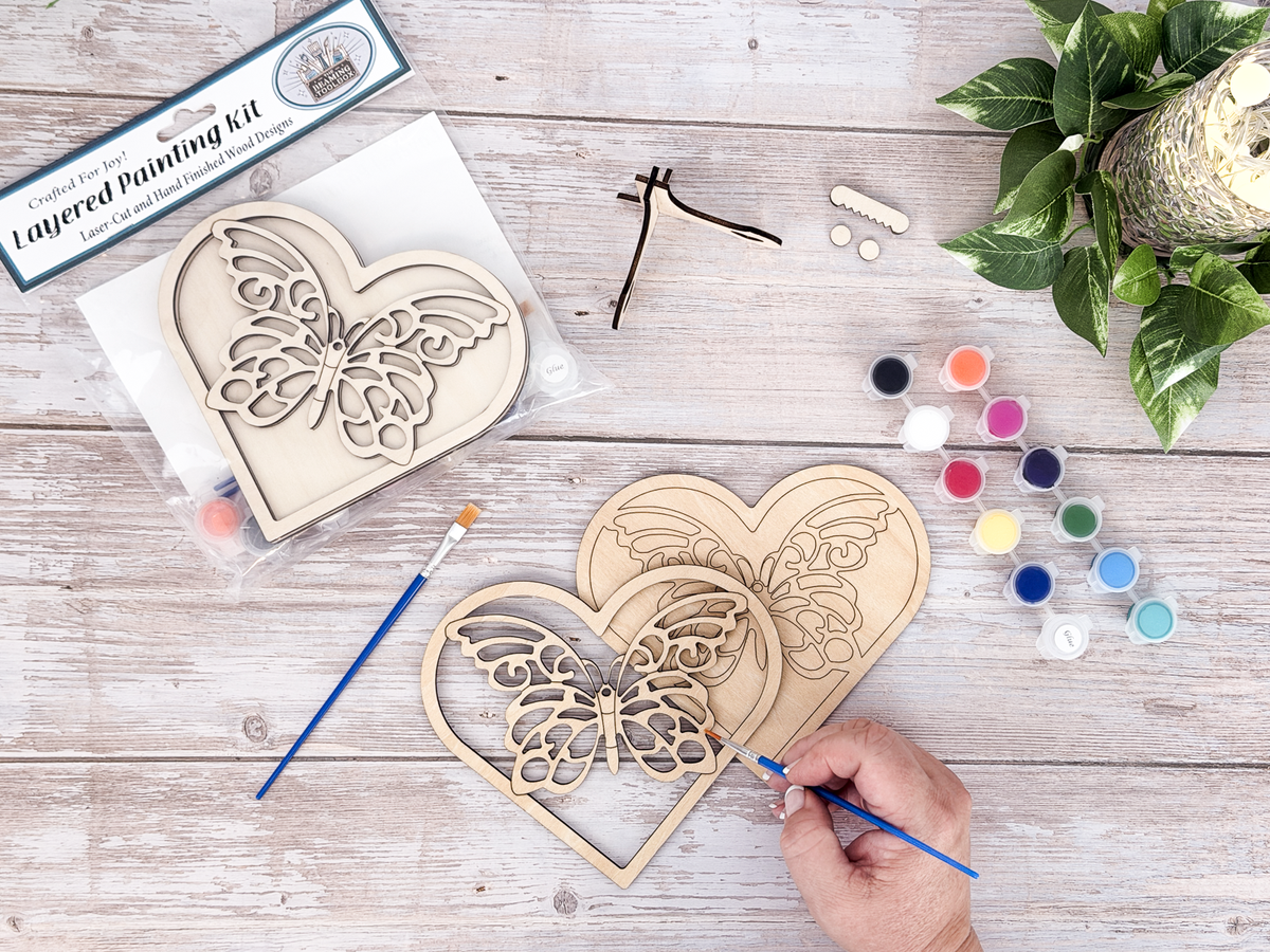 Person painting the butterfly heart layered wood craft kit on a rustic wood background with paint pots, brushes, and greenery.