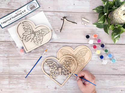 Person painting the butterfly heart layered wood craft kit on a rustic wood background with paint pots, brushes, and greenery.