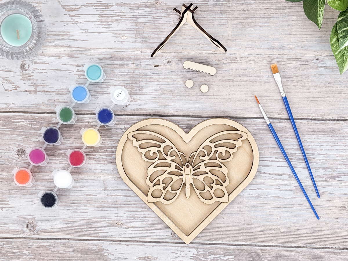 Unpainted butterfly heart layered wood artwork displayed with paint pots, brushes, stand pieces, candle, and greenery.