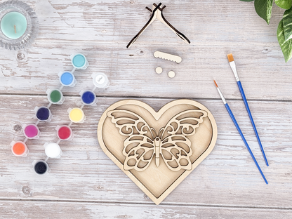 Unpainted butterfly heart layered wood artwork displayed with paint pots, brushes, stand pieces, candle, and greenery.