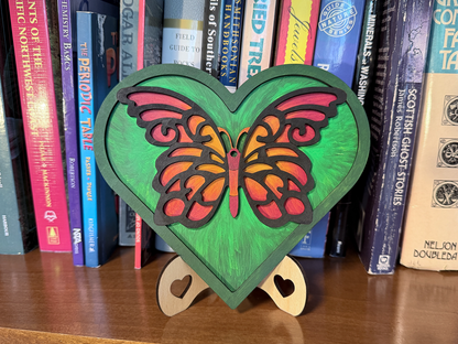 A heart-shaped wooden wall decor piece featuring a colorful butterfly design, displayed on a wooden shelf in front of a row of books.