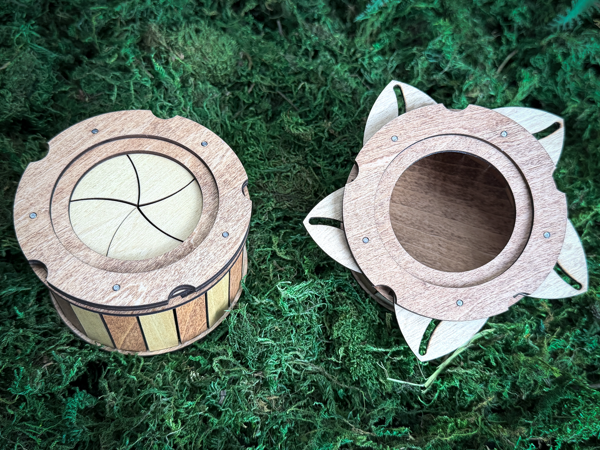 Two circular wooden containers with mechanical iris lids. One container is closed, showing a light-colored iris mechanism. The other container is open, revealing a hollow interior and petal-like wooden extensions around the rim.