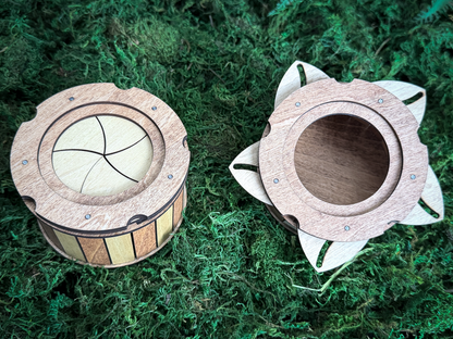 Two circular wooden containers with mechanical iris lids. One container is closed, showing a light-colored iris mechanism. The other container is open, revealing a hollow interior and petal-like wooden extensions around the rim.
