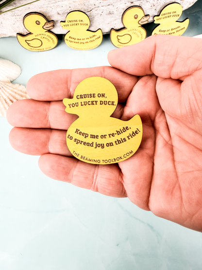 Hand holding the back of a wooden cruise duck token engraved with a cruise hiding message and website text.