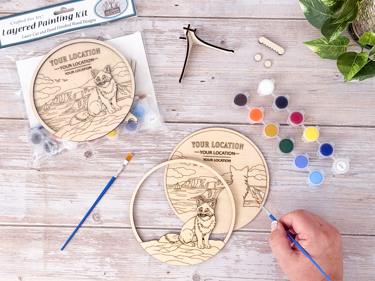 Person painting the Custom Location Fox layered wood craft kit on a rustic background with paint pots, brushes, packaging, and display hardware.