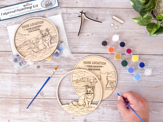 Person painting the Custom Location Fox layered wood craft kit on a rustic background with paint pots, brushes, packaging, and display hardware.