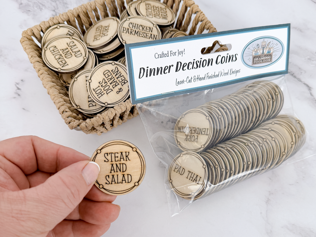 A hand holds a round wooden coin engraved with 'STEAK AND SALAD' in front of a basket and clear bag of matching wooden dinner decision coins on a white marble surface. The package label reads 'Crafted For Joy! Dinner Decision Coins' and shows multiple engraved coin designs.